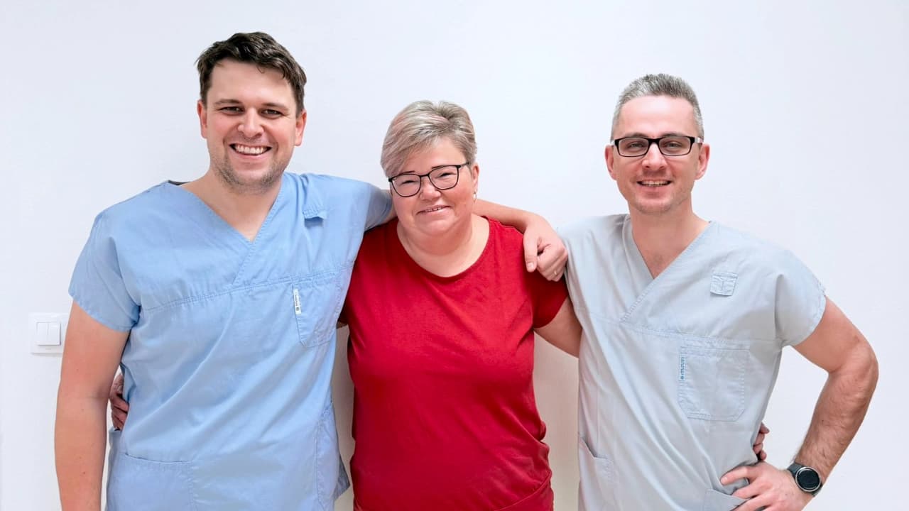 Tři usmívající se zdravotníci v modrém, červeném a šedém oděvu stojí v řadě u bílé zdi a drží se kolem ramen.